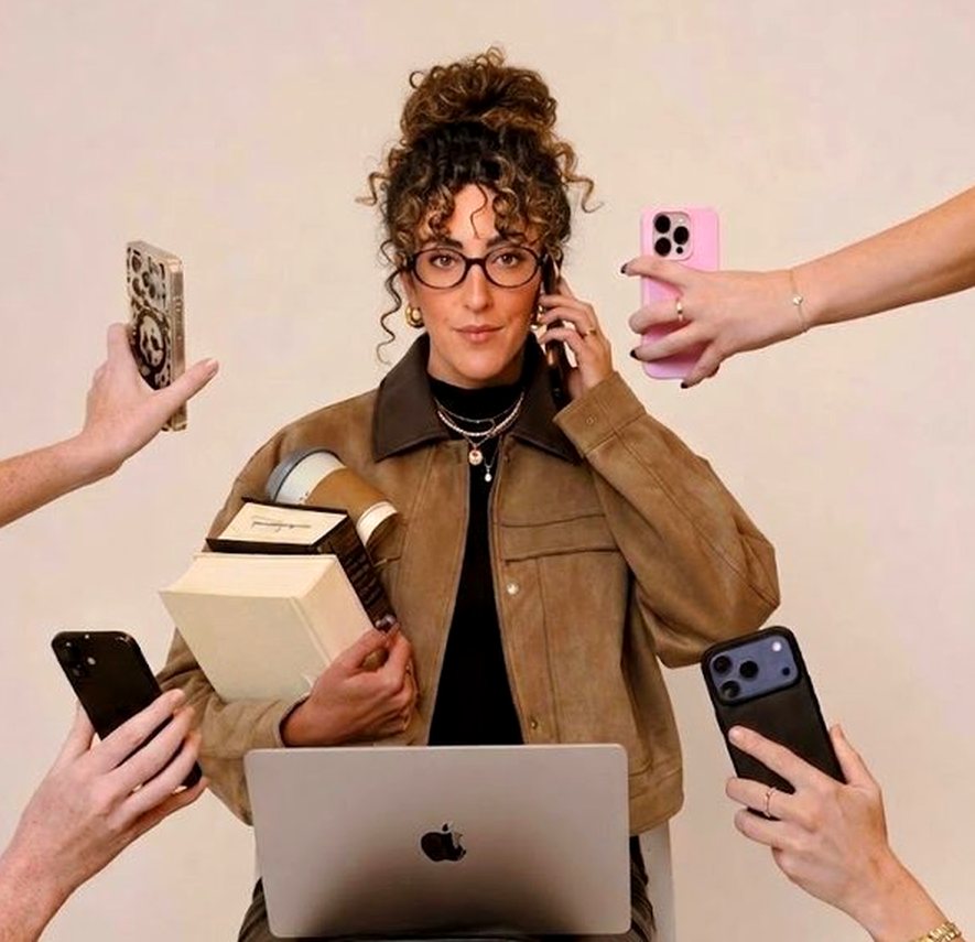 Marta Vannelli seduta con il computer sulle ginocchia, i libri in braccio e un telefono all’orecchio, mentre da ogni lato delle mani le porgono altri telefoni